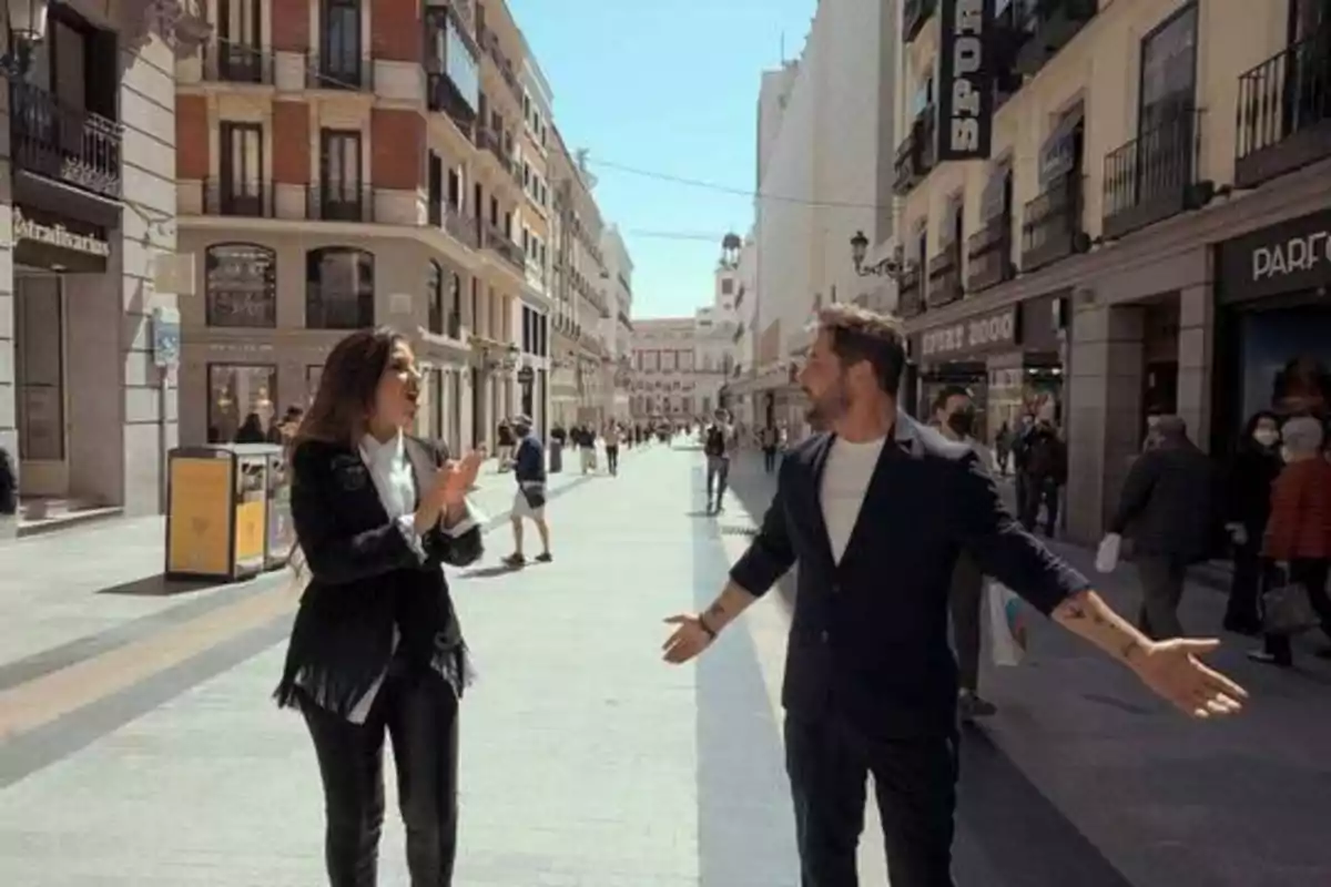 Dos personas conversando en una calle peatonal rodeada de edificios y tiendas.