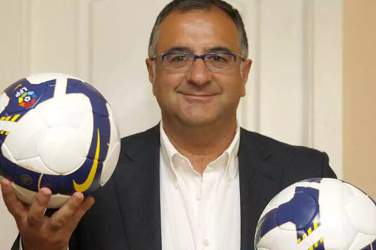 Hombre con gafas sosteniendo dos balones de fútbol.