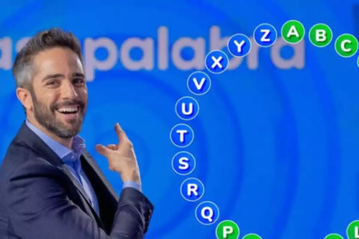 Un hombre sonriente con barba y traje oscuro señala hacia un conjunto de letras en un fondo azul que forman un semicírculo, con la palabra "palabra" parcialmente visible en el fondo.