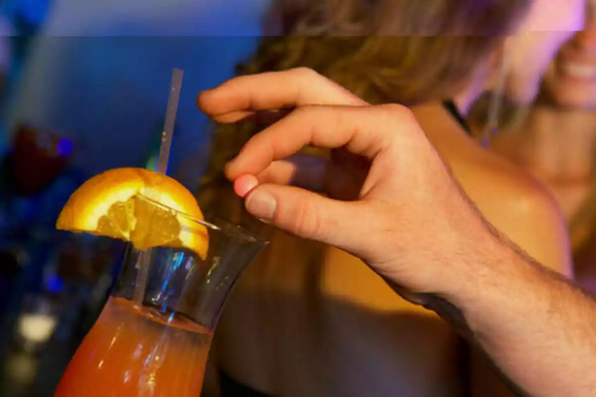 Una mano sosteniendo una pastilla sobre un vaso de cóctel adornado con una rodaja de naranja.