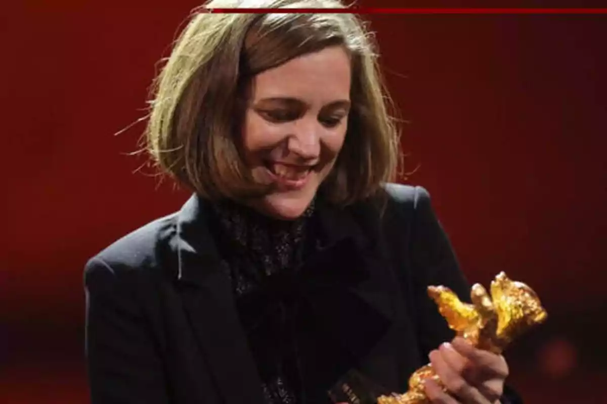 Persona sonriendo mientras sostiene un trofeo dorado en un fondo rojo.