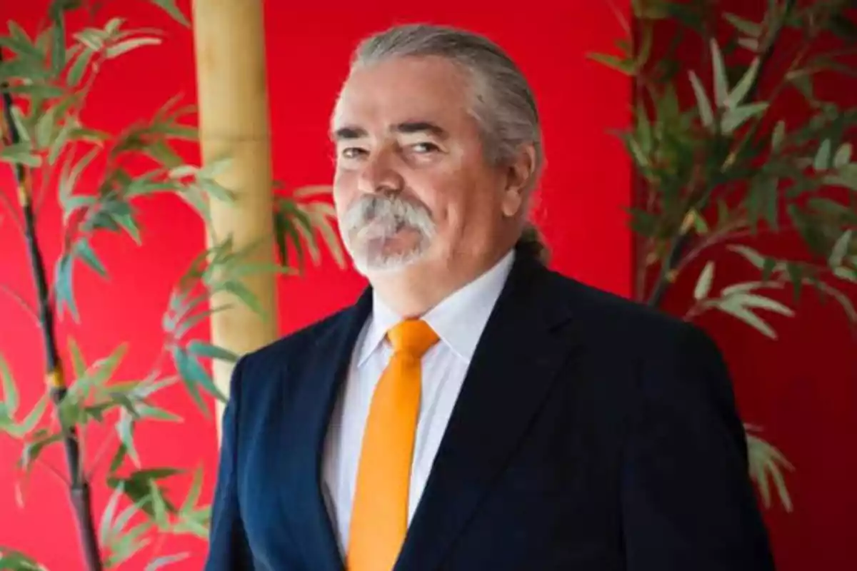 Hombre de cabello canoso y barba, vestido con traje oscuro y corbata naranja, posando frente a un fondo rojo con plantas.