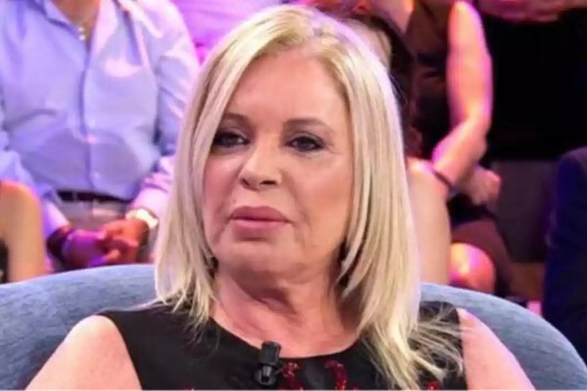 Una mujer rubia con cabello lacio y expresión seria, sentada en un programa de televisión con personas en el fondo.