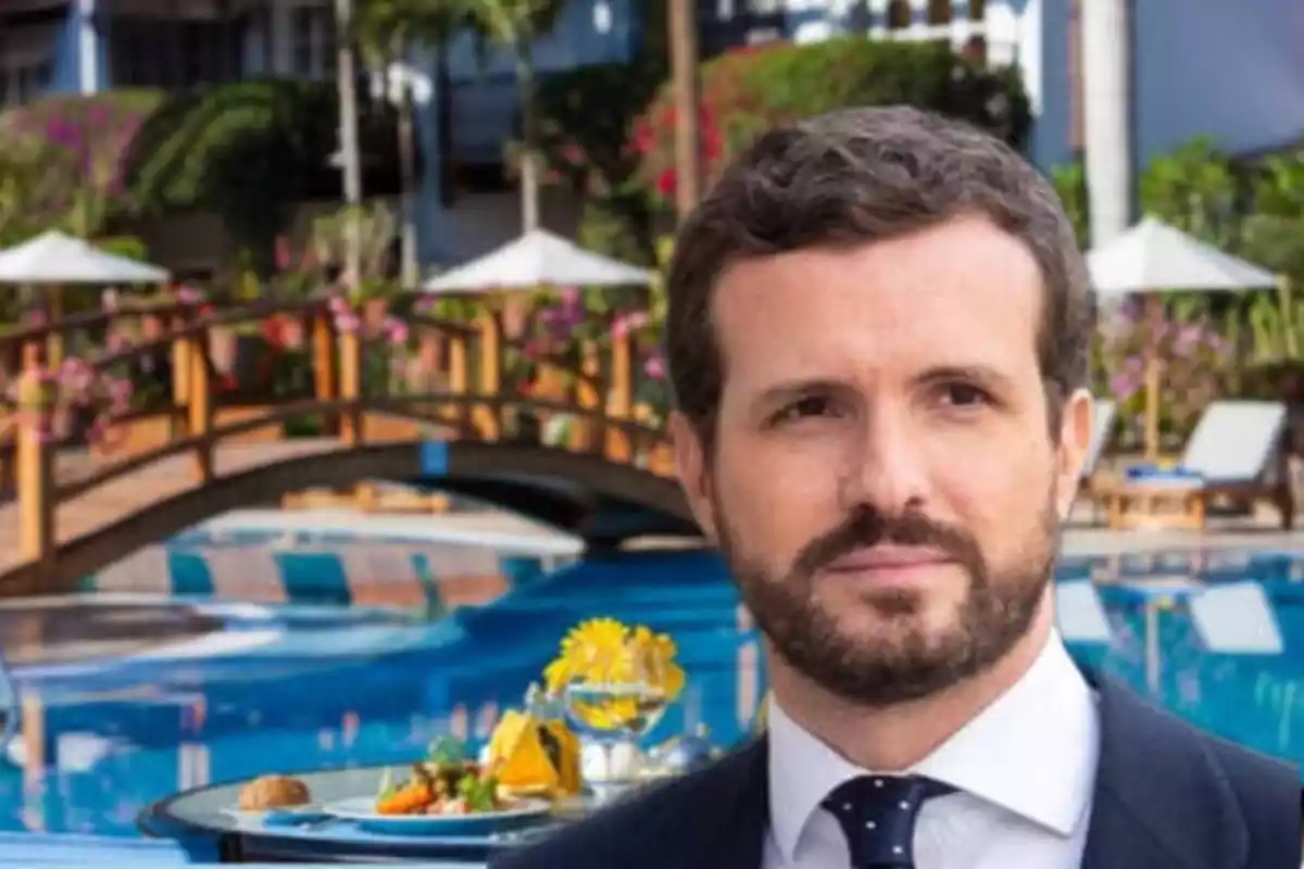 Un hombre con barba y traje está frente a una piscina con un puente de madera y flores en el fondo.