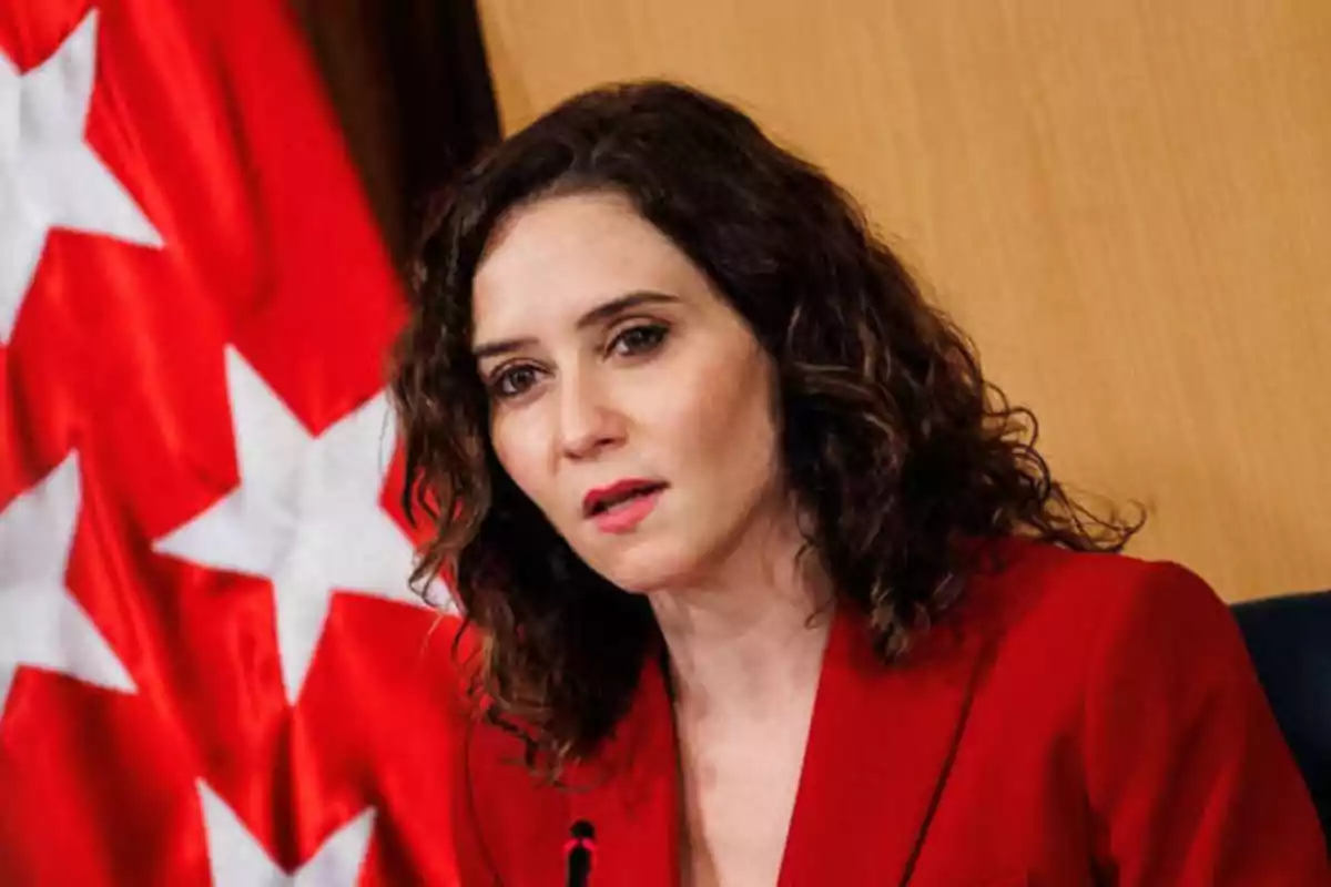 Mujer con chaqueta roja hablando frente a una bandera roja con estrellas blancas.