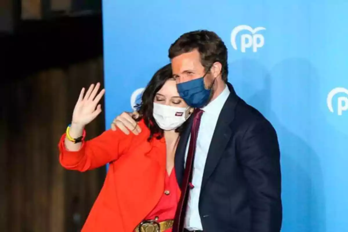 Dos personas con mascarillas, una de ellas saludando, frente a un fondo azul con el logotipo del Partido Popular.