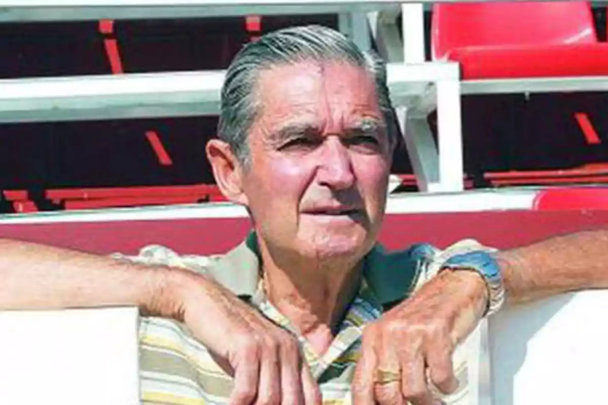 Hombre mayor con camisa de rayas apoyado en una barandilla en un estadio con asientos rojos.