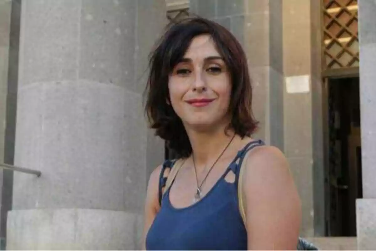 Una mujer con cabello corto y oscuro, vestida con una camiseta sin mangas azul, está de pie frente a un edificio de piedra.