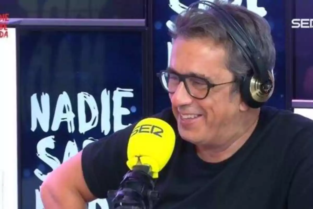 Un hombre con gafas y auriculares hablando en un micrófono amarillo con la palabra "SER" en un estudio de radio con un cartel de fondo que dice "NADIE".