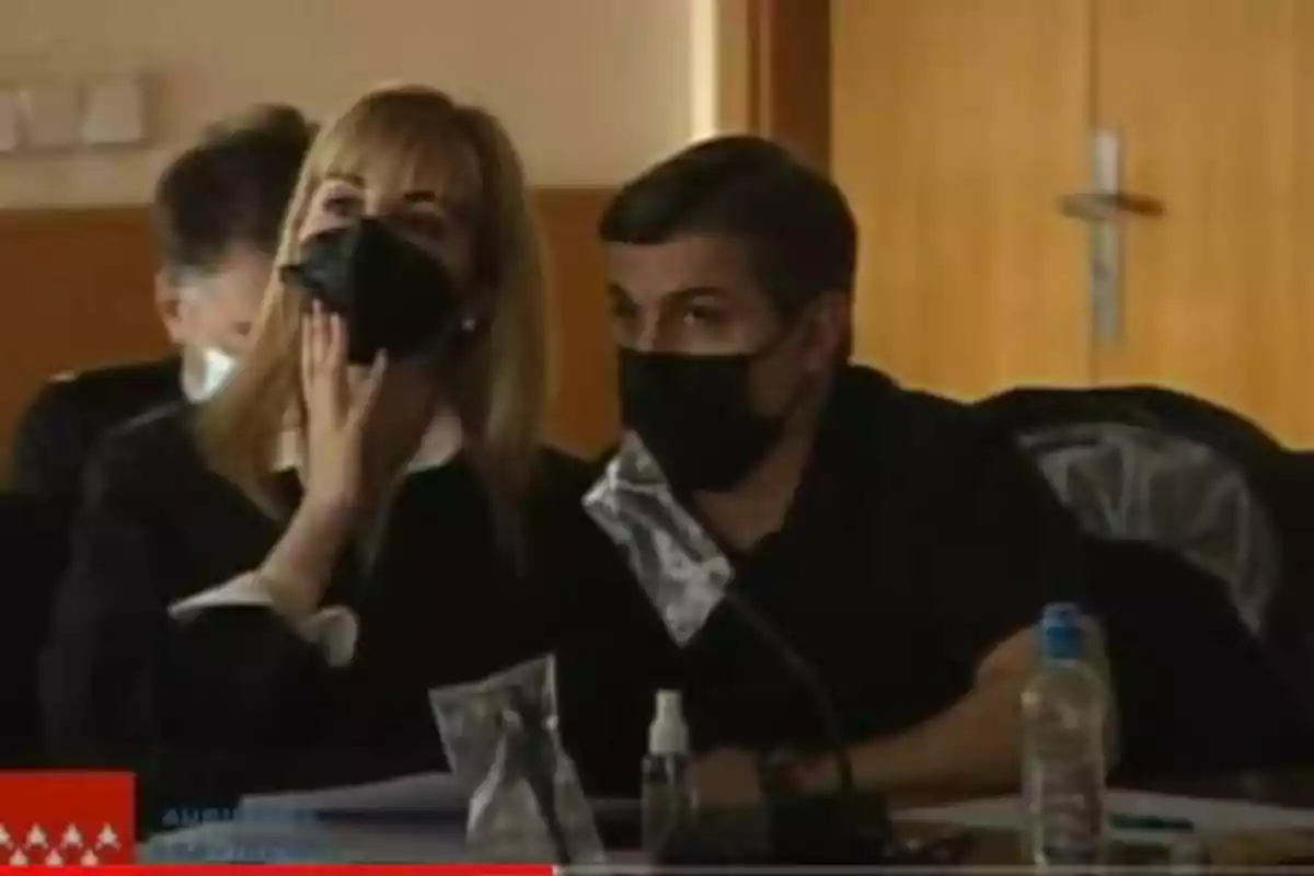 Dos personas con mascarillas negras están sentadas en una mesa, una de ellas tocándose la cara, en un entorno que parece ser una sala de reuniones o conferencia.