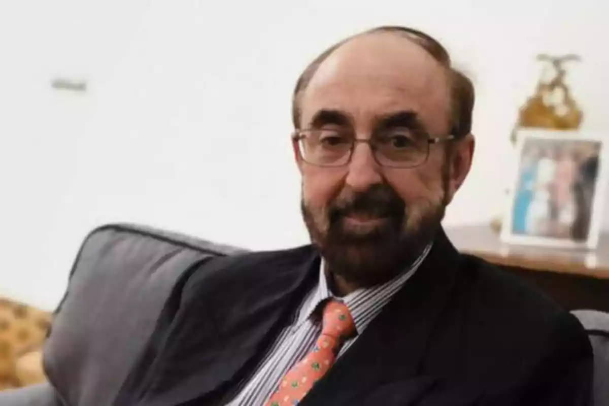 Hombre con gafas y barba sentado en un sofá con una corbata naranja y una camisa a rayas.