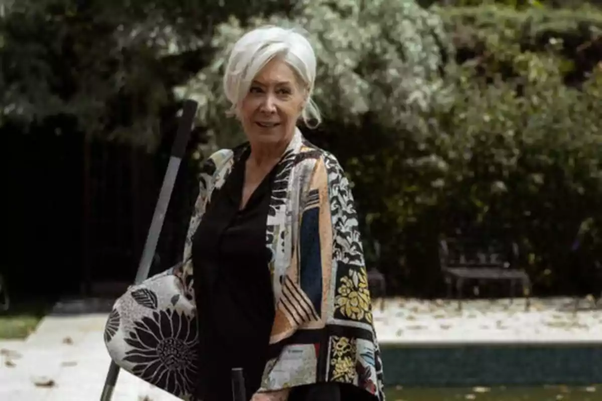 Mujer mayor con cabello blanco y ropa colorida, de pie al aire libre junto a una piscina.