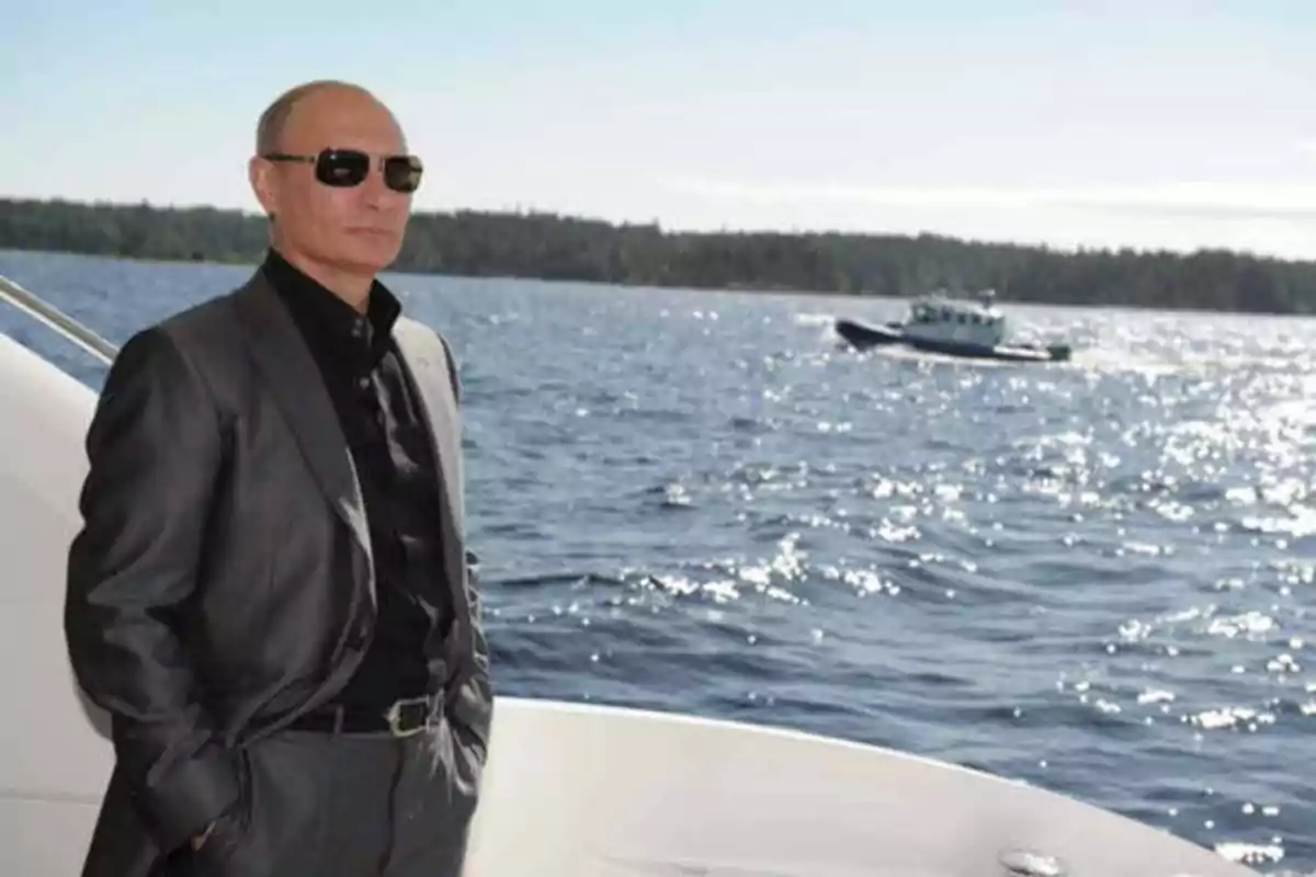 Hombre con traje oscuro y gafas de sol de pie en un barco con el mar y un bote en el fondo.