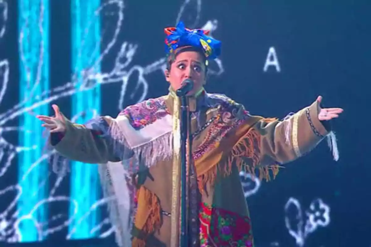 Persona cantando en un escenario con un atuendo colorido y un tocado en la cabeza.