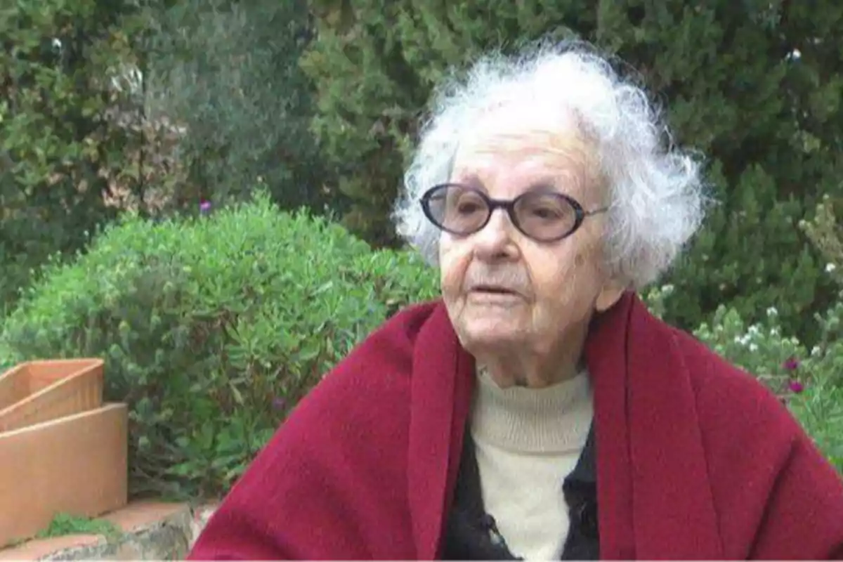 Una anciana con gafas y cabello canoso, envuelta en una manta roja, está sentada al aire libre con vegetación de fondo.