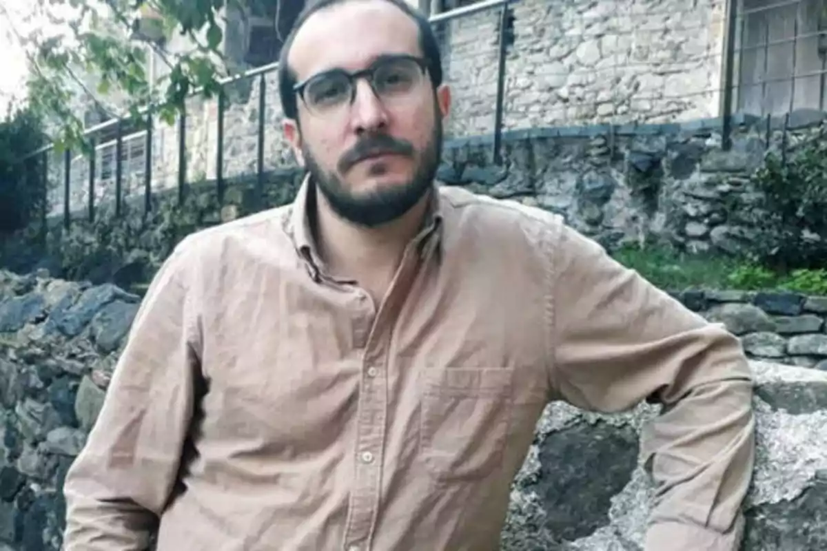 Hombre con gafas y barba, vestido con una camisa beige, posando frente a una pared de piedra.