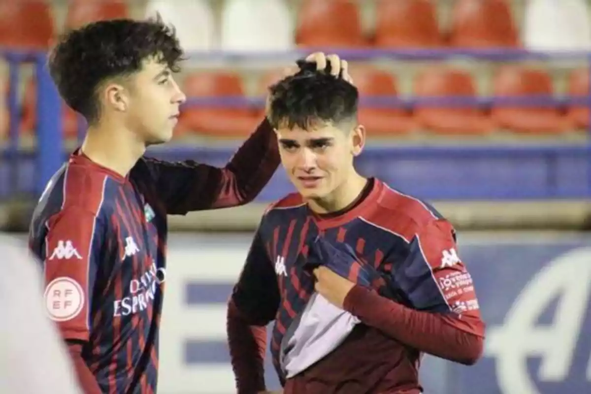 Dos jugadores de fútbol con uniformes rojos y azules, uno de ellos consuela al otro que parece estar triste.