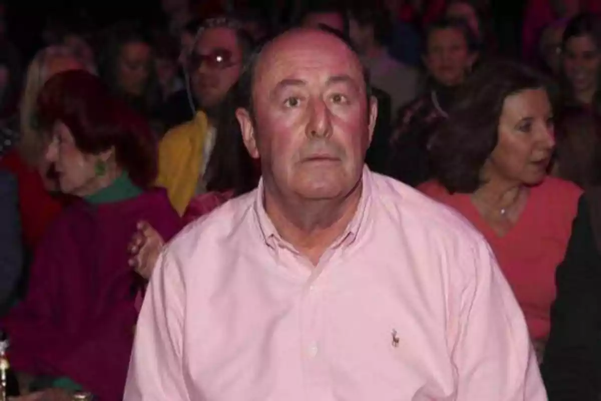 Un hombre con camisa rosa está sentado en un lugar lleno de gente.