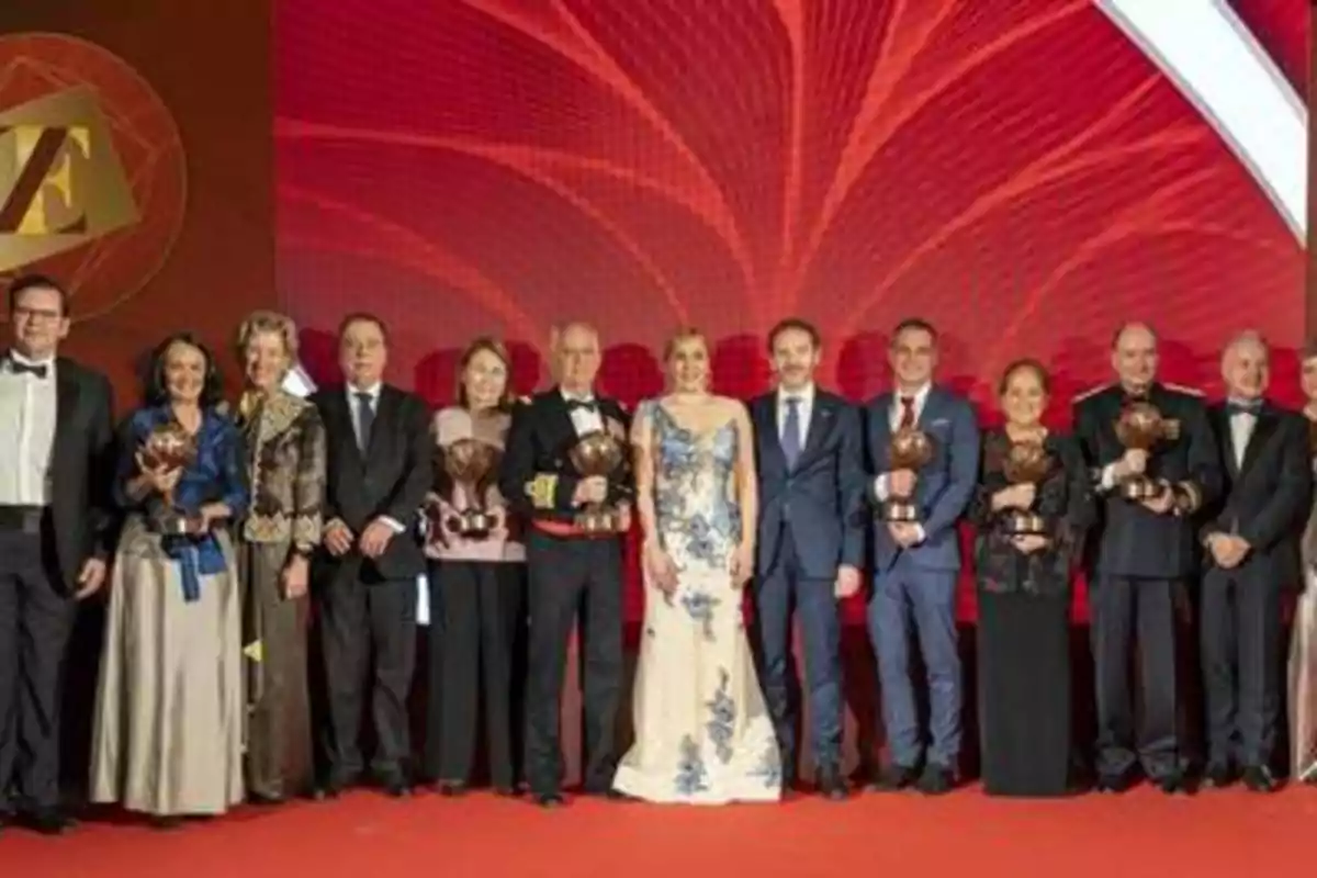 Un grupo de personas posando en un evento formal, algunas de ellas sosteniendo trofeos, con un fondo rojo decorativo.