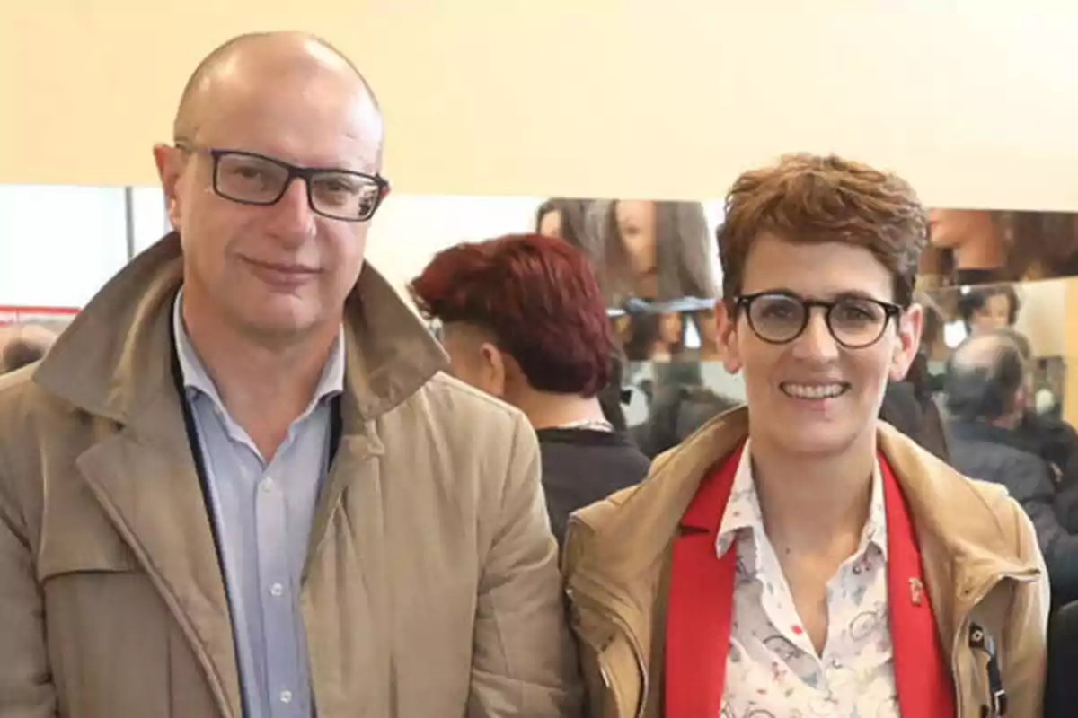 Dos personas posan juntas, ambas llevan gafas y abrigos claros, una de ellas tiene el cabello corto y la otra es calva.