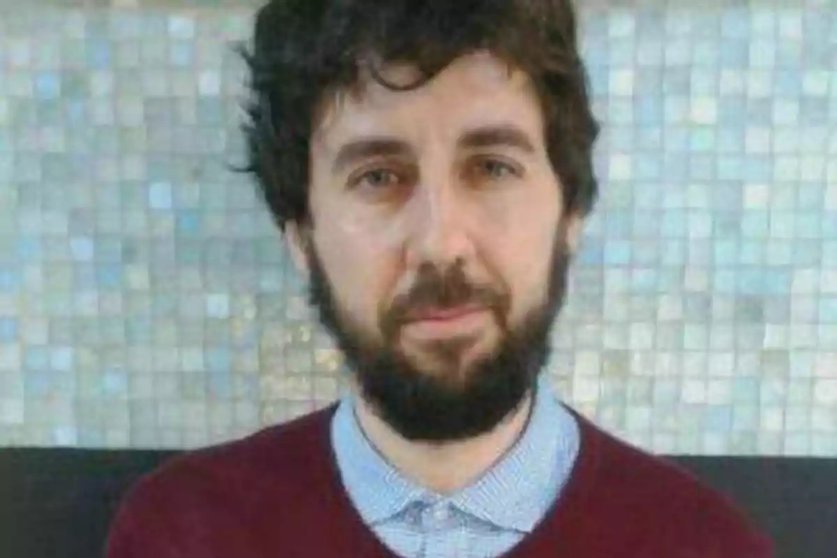 Hombre con barba y cabello oscuro, vistiendo un suéter rojo sobre una camisa azul claro, posando frente a un fondo de mosaicos.