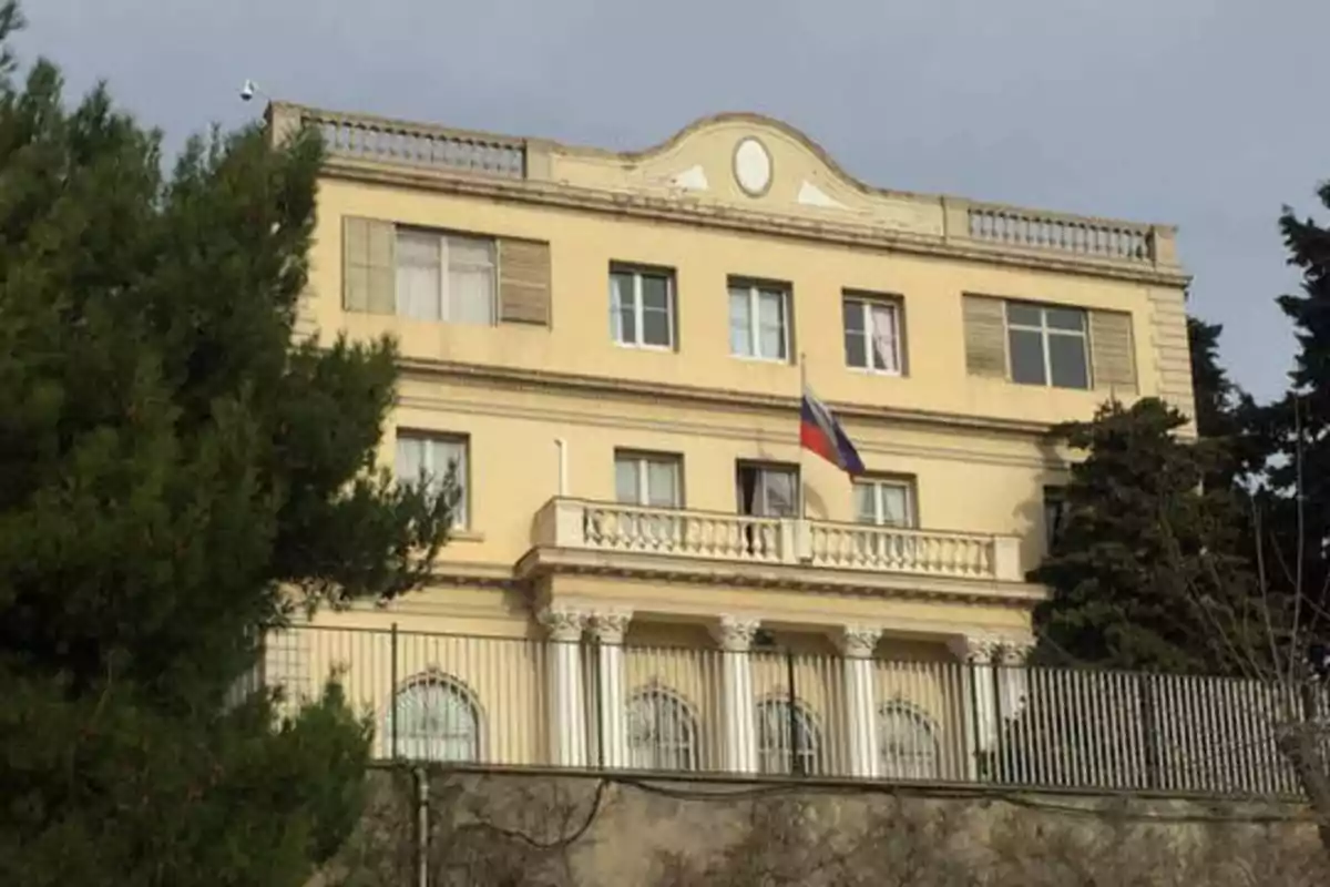 Edificio de tres pisos con una bandera en el balcón y árboles alrededor.