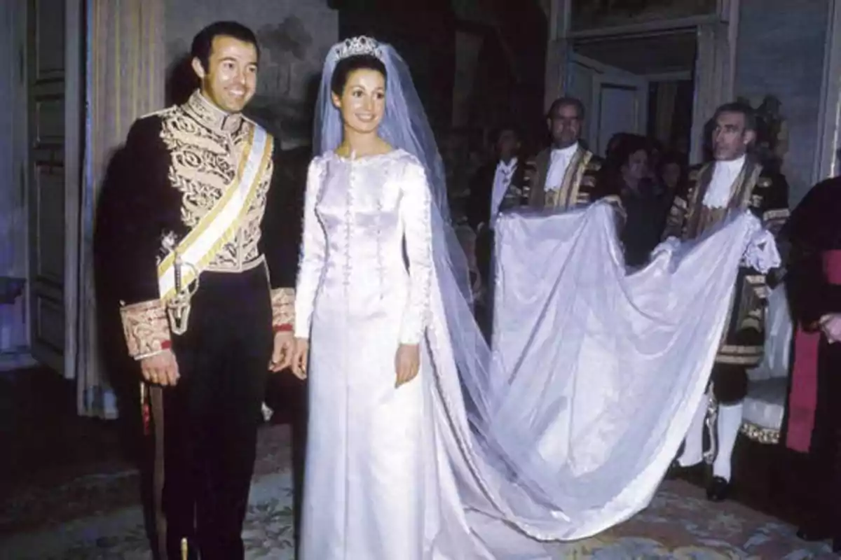 Una pareja vestida de gala en su boda, con la novia usando un vestido blanco largo y un velo, y el novio con un uniforme formal, acompañados por asistentes que sostienen la cola del vestido.