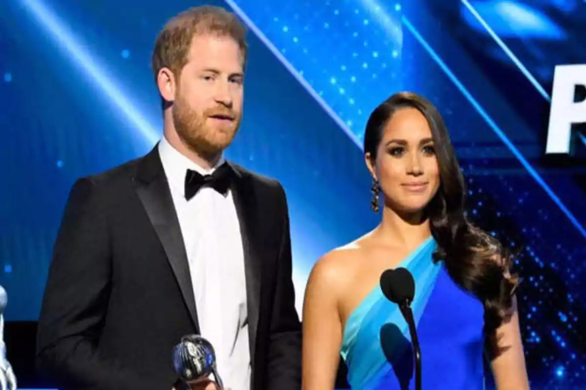 Una pareja elegantemente vestida en un escenario con fondo azul y luces brillantes, él con un esmoquin negro y ella con un vestido azul de un solo hombro.