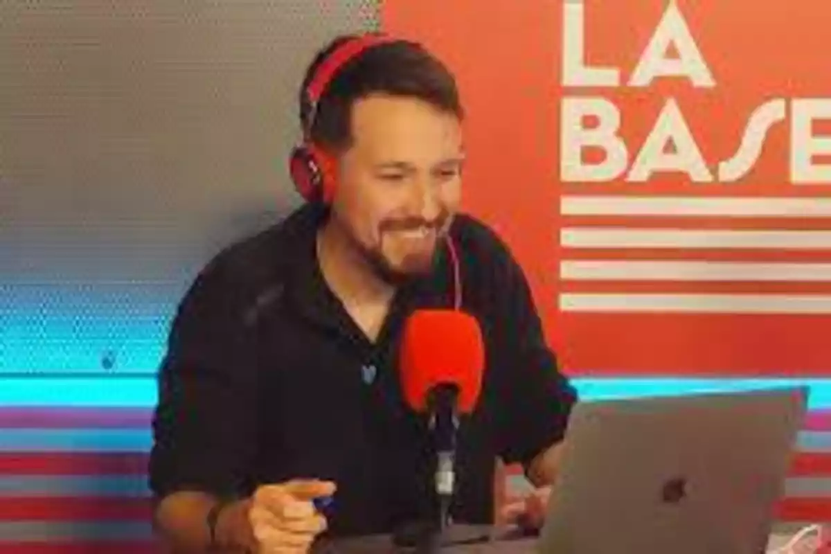 Un hombre con auriculares rojos y un micrófono rojo frente a una computadora portátil, con un fondo rojo que tiene el texto "LA BASE".