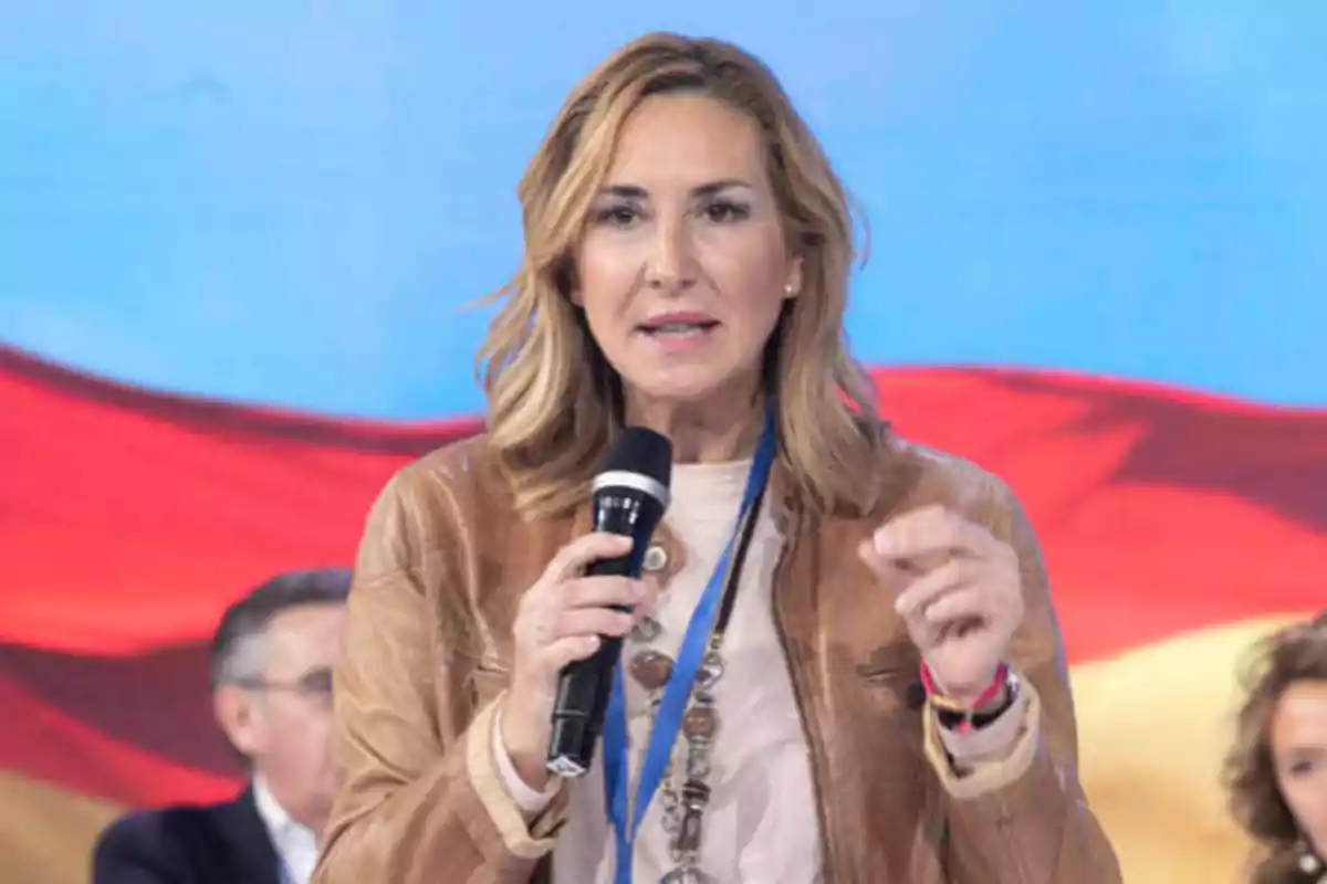 Mujer hablando en un evento con un micrófono en la mano y una bandera de fondo.