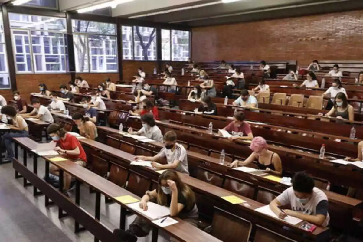 Estudiantes en un aula grande tomando un examen, sentados en escritorios de madera con distanciamiento social y usando mascarillas.