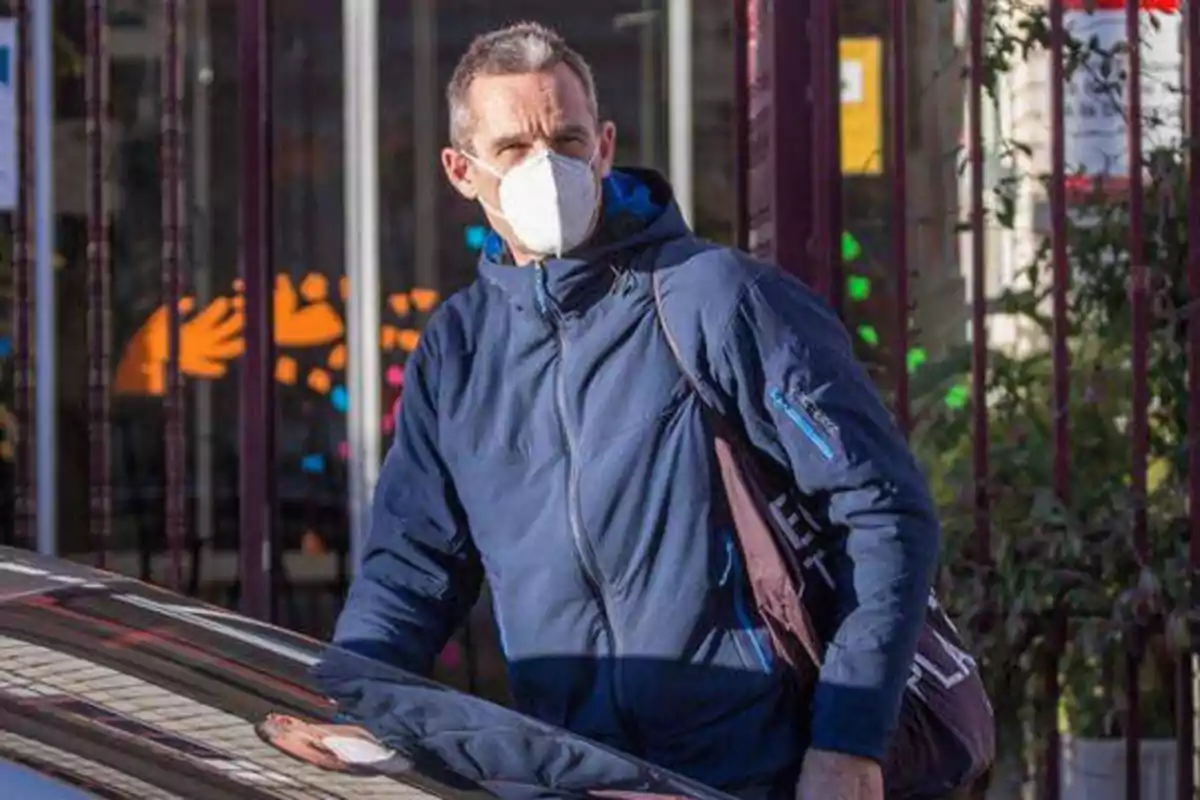 Hombre con chaqueta azul y mascarilla blanca, de pie junto a un coche en una calle.