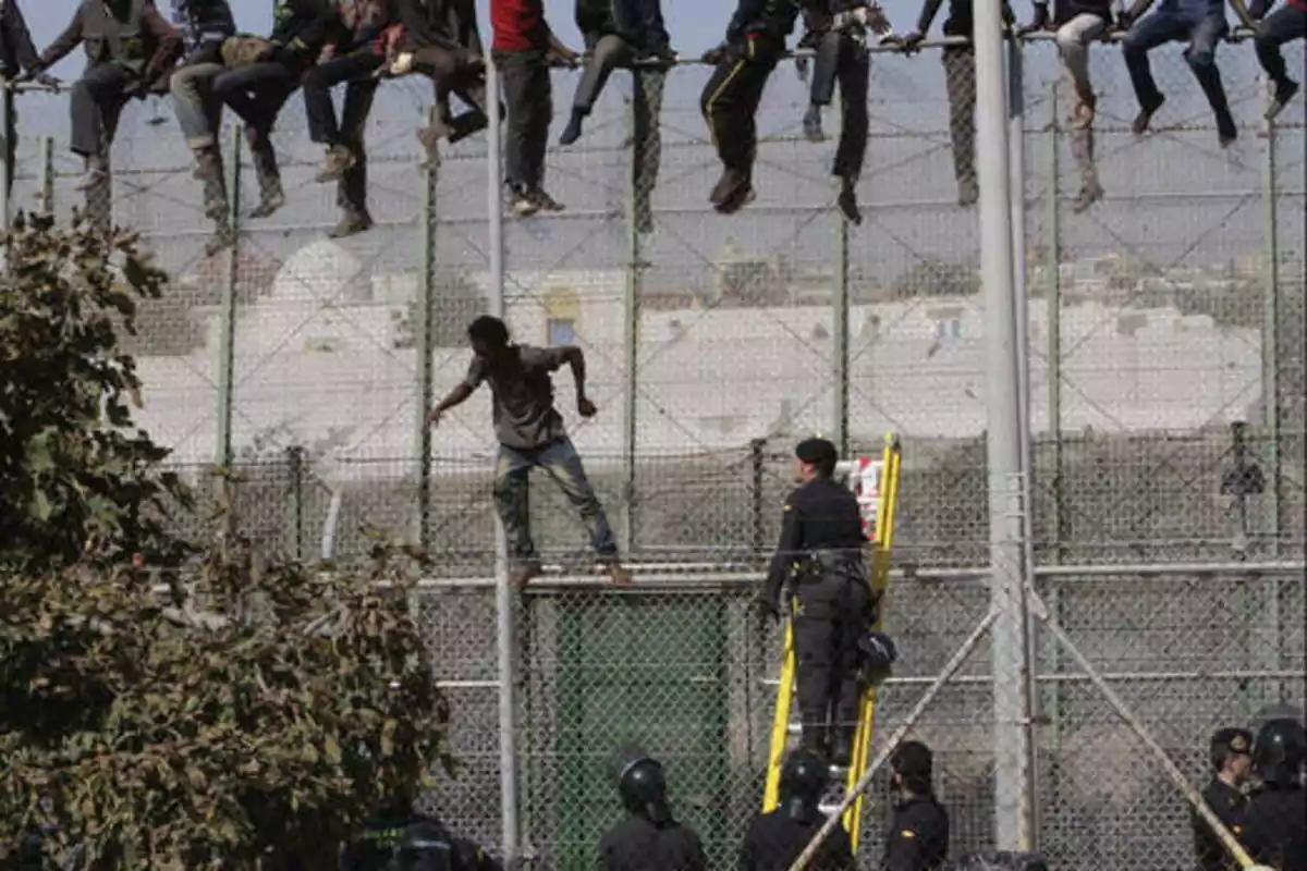 Un grupo de personas se encuentra sentado en la parte superior de una valla alta, mientras que una persona está en medio de la escalada y varios agentes de seguridad observan desde abajo con una escalera apoyada en la valla.