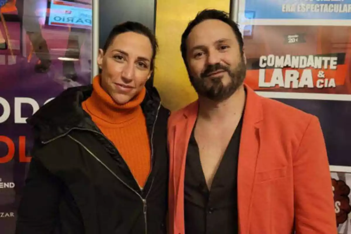 Dos personas posando frente a un cartel publicitario, una mujer con suéter naranja y chaqueta negra y un hombre con chaqueta roja y camisa negra.