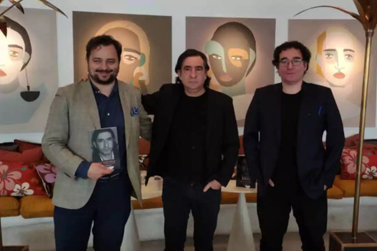 Tres hombres posan juntos frente a una pared con pinturas abstractas, uno de ellos sostiene un libro con una fotografía en blanco y negro en la portada.