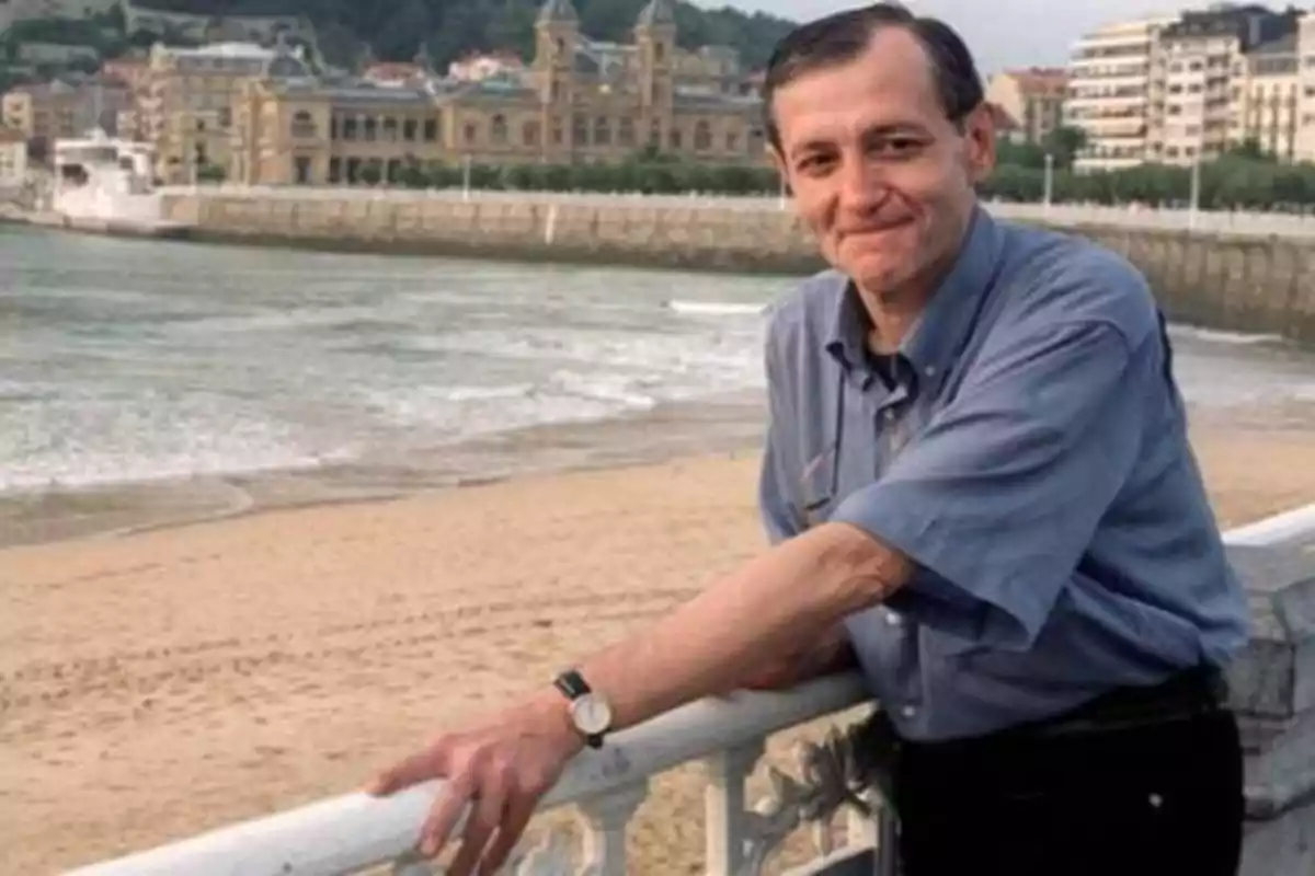 Un hombre sonriente con camisa azul está apoyado en una barandilla frente a una playa con edificios y montañas al fondo.