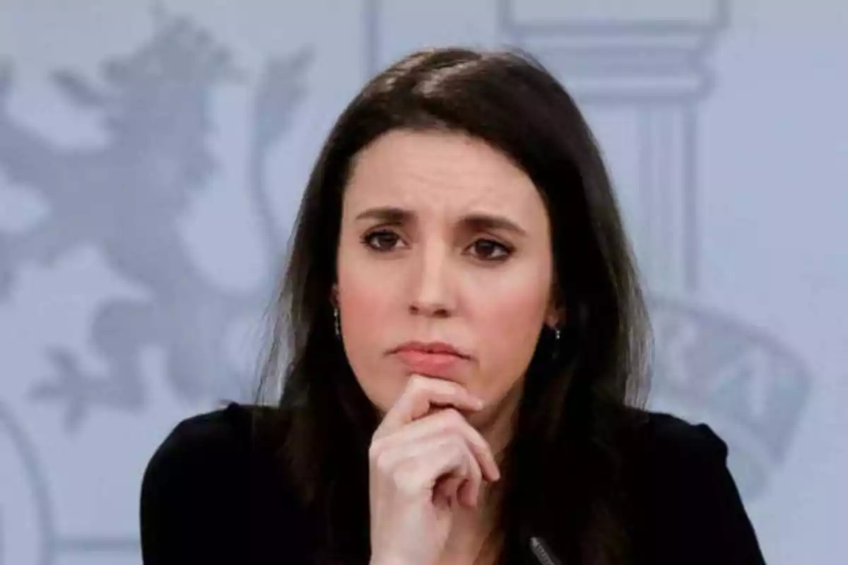 Mujer con cabello oscuro y expresión pensativa, con la mano en el mentón, frente a un fondo gris con un diseño borroso.