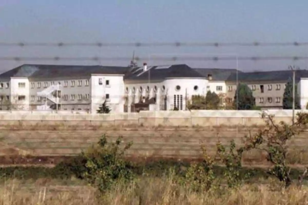 Edificio grande de varios pisos con ventanas, rodeado por una cerca de alambre de púas y un muro, con vegetación en primer plano.