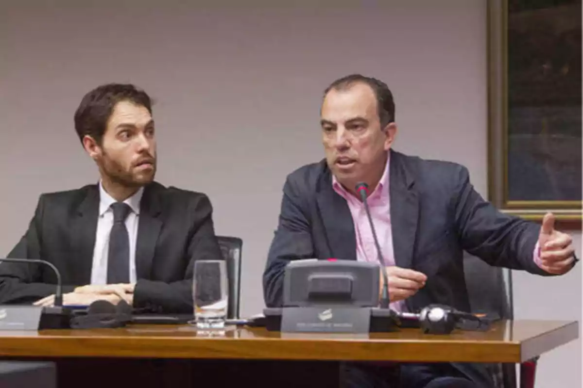 Dos hombres en una reunión, uno de ellos hablando y gesticulando mientras el otro lo observa.