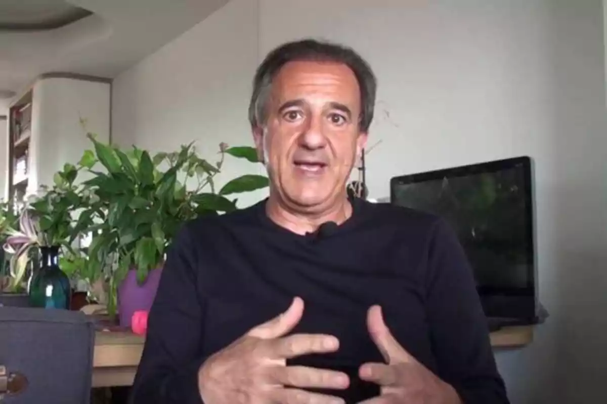 Un hombre de mediana edad con una camiseta negra está sentado frente a una mesa con plantas y una computadora portátil en el fondo.