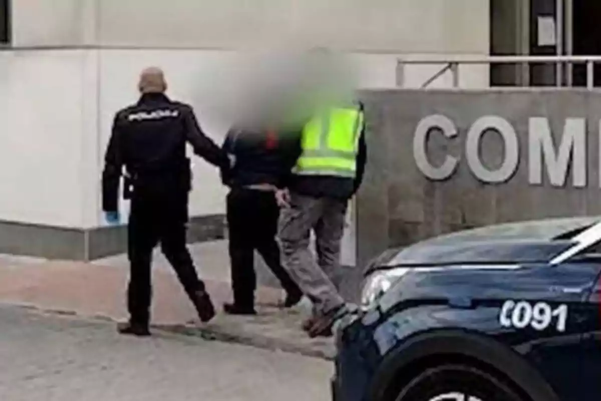 Dos agentes de policía escoltan a una persona con el rostro difuminado hacia un edificio con la palabra "COMI" visible en la pared, junto a un coche patrulla con el número "091".