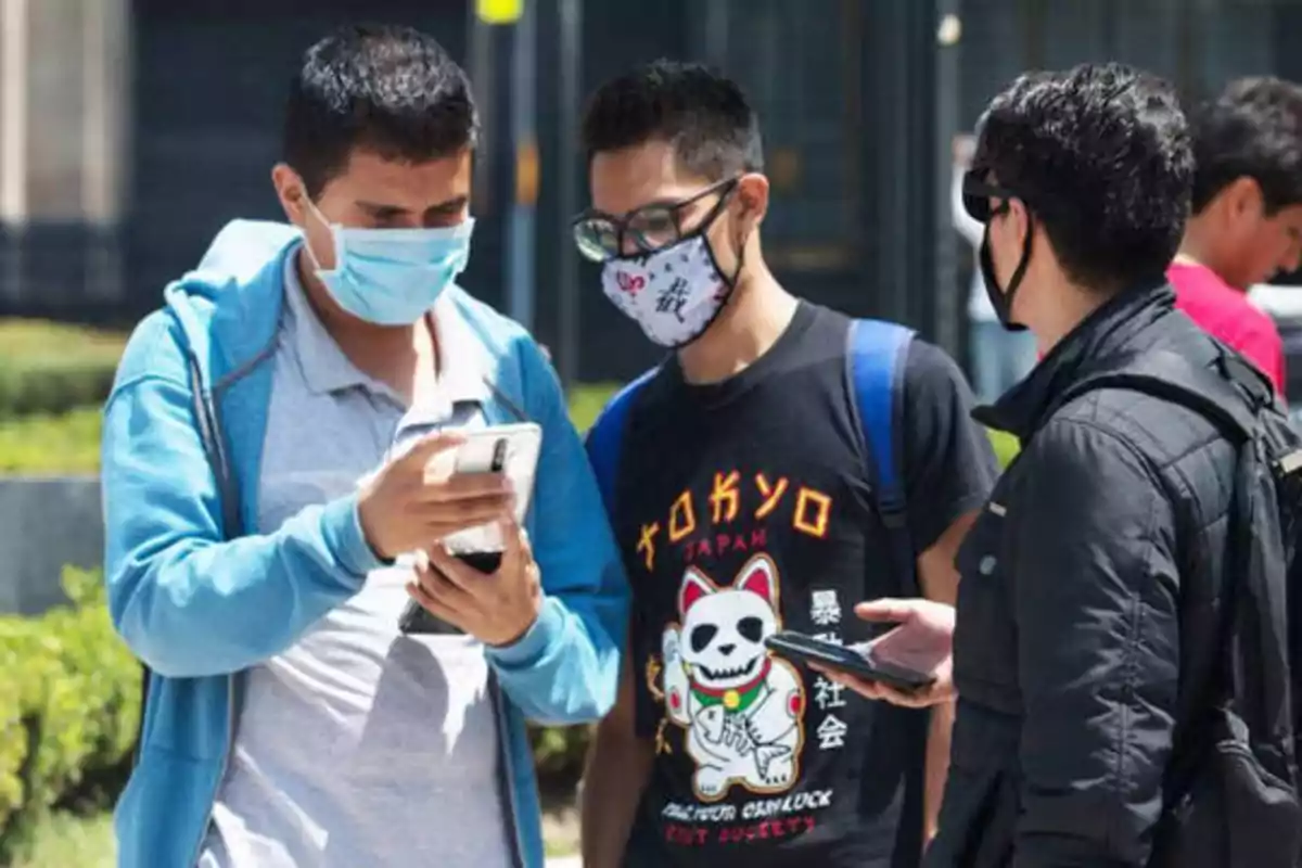 Tres personas con mascarillas conversan mientras miran sus teléfonos móviles en un espacio al aire libre.