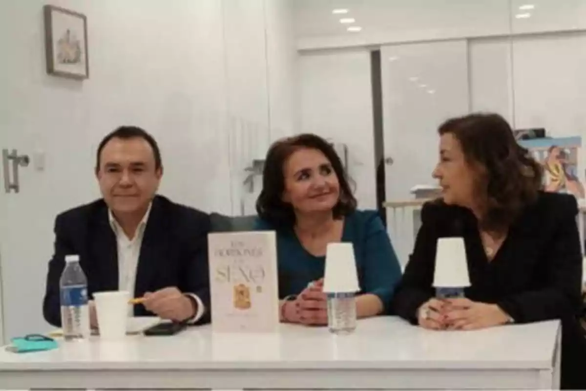 Tres personas sentadas en una mesa con una botella de agua, vasos y un libro en el centro.