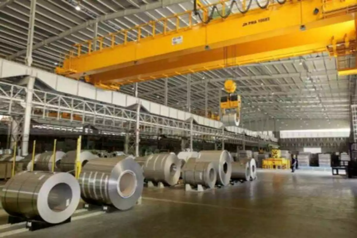 Interior de una fábrica con bobinas de metal y una grúa amarilla.