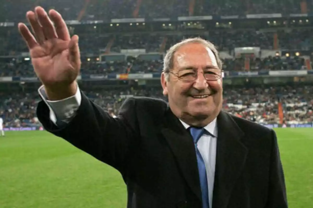 Un hombre mayor con gafas y traje oscuro saluda con la mano levantada en un estadio de fútbol lleno de espectadores.