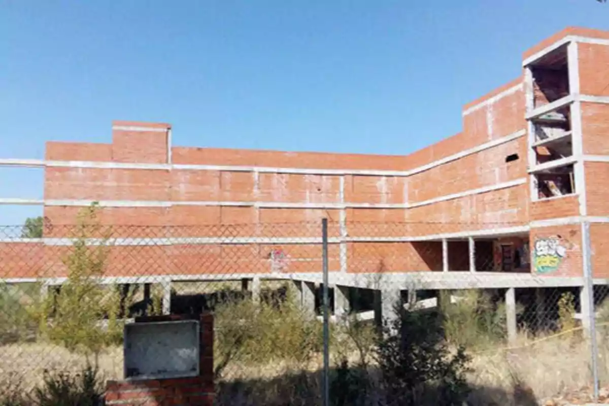 Edificio de ladrillo sin terminar rodeado por una cerca de alambre bajo un cielo despejado.