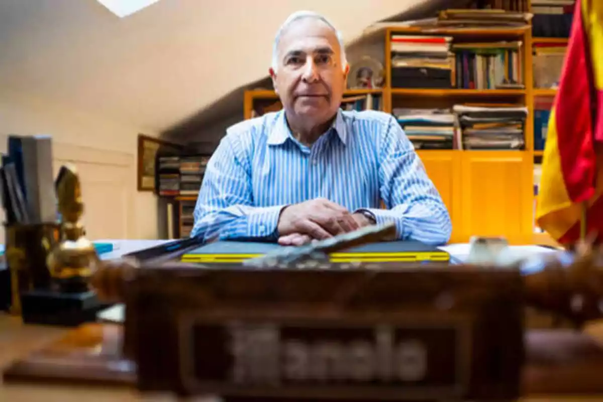 Hombre mayor sentado en un escritorio con estanterías llenas de libros al fondo y una bandera a la derecha.