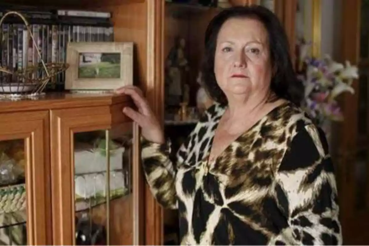 Una mujer de cabello oscuro y expresión seria, vestida con una blusa de estampado animal, está de pie junto a un mueble de madera con puertas de vidrio, sobre el cual hay una canasta metálica y un marco de fotos.