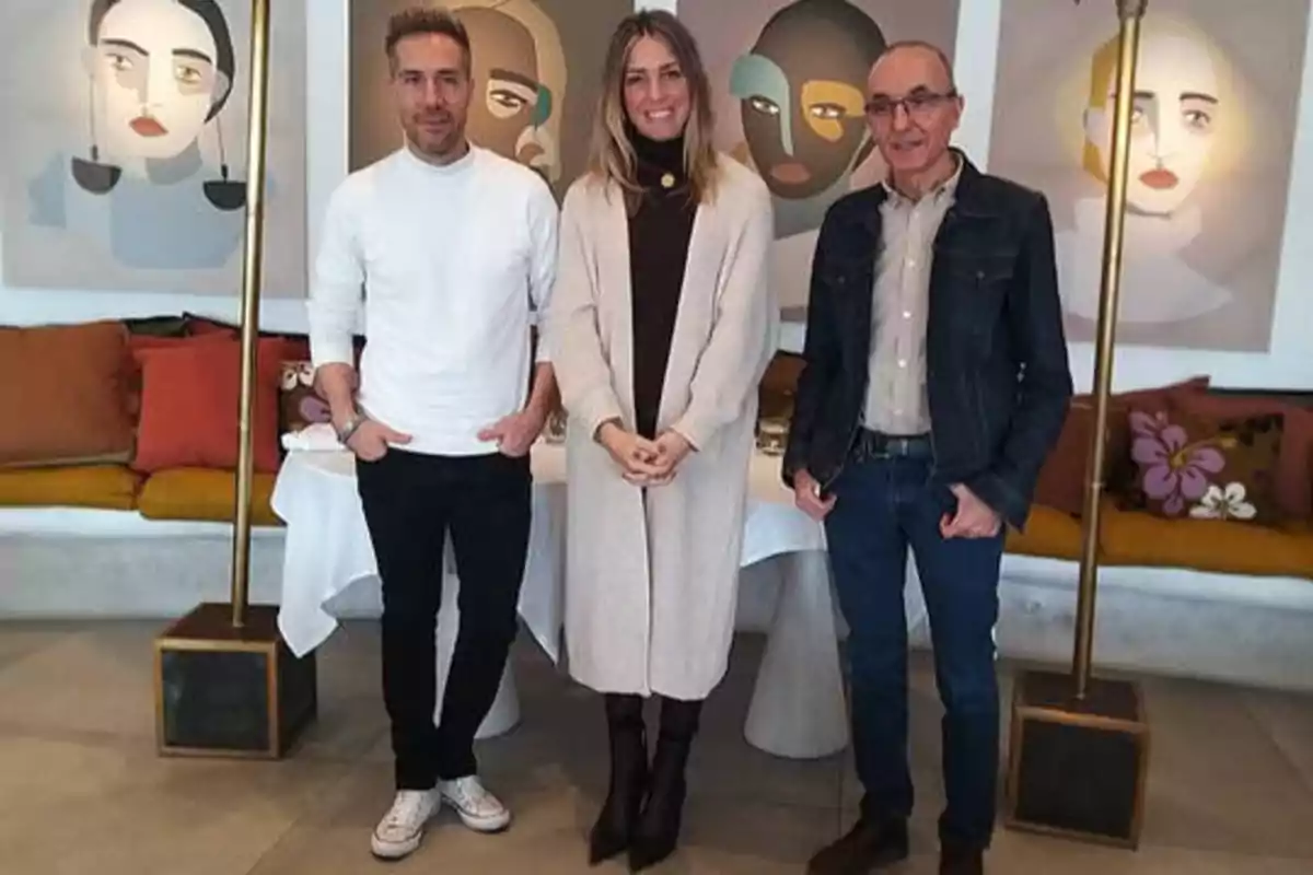 Tres personas posan juntas frente a una mesa, con cuadros de rostros abstractos en la pared detrás de ellos.