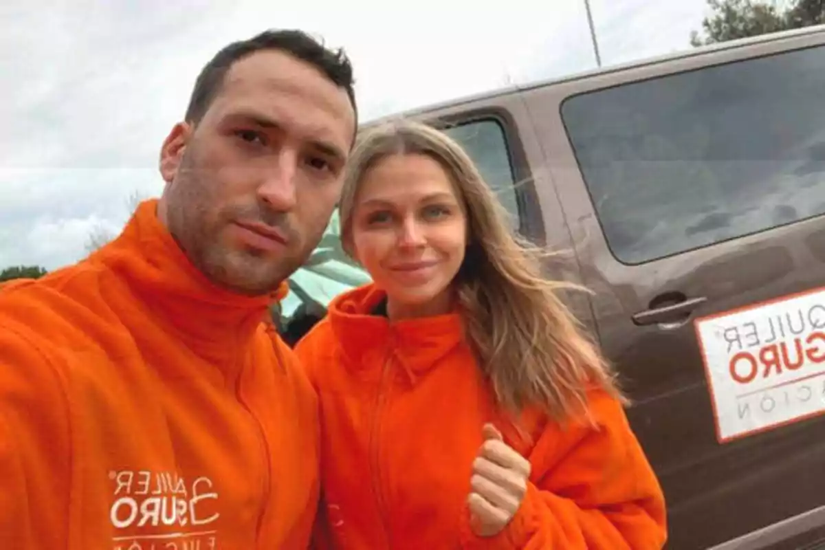 Dos personas con chaquetas naranjas posan para una selfie frente a una camioneta marrón con un letrero en la puerta.